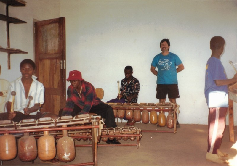 Ted standing in a room at Cyrene school with African boys playing xylophones.